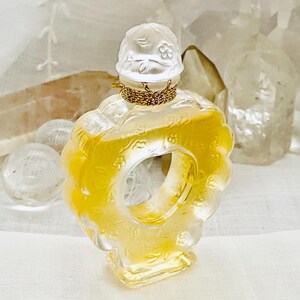 May include: A clear glass bottle of perfume with a gold cap and a floral design. The bottle is shaped like a ring and has a yellow liquid inside.