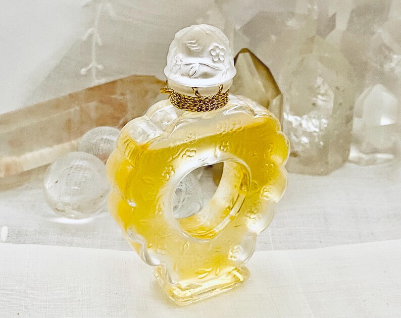 May include: A clear glass bottle of perfume with a gold cap and a floral design. The bottle is shaped like a ring and has a yellow liquid inside.