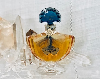 May include: A vintage bottle of Shalimar perfume by Guerlain. The bottle is clear glass with a gold-colored liquid inside. The bottle has a unique, ornate design with a fan-shaped stopper. The stopper is blue and has the words "Shalimar Parfum" written on it.