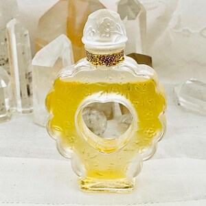 May include: A heart-shaped glass bottle of perfume with a white cap and gold accents. The bottle is filled with a light yellow liquid.
