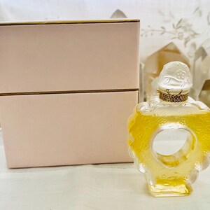 May include: A heart-shaped glass bottle of perfume with a gold stopper and a floral design. The bottle is sitting on a pink box with a gold trim.
