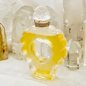May include: A clear glass bottle of perfume with a gold cap and a floral design. The bottle is filled with a yellow liquid and is sitting on a white surface.