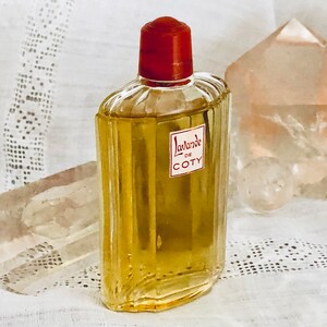 Pu&ograve; includere: Un flacone di profumo Coty L'Lavande in vetro trasparente con un tappo rosso. Il flacone &egrave; appoggiato su un tovagliolo di pizzo bianco.
