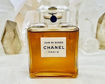 May include: A rectangular glass perfume bottle filled with amber liquid. The label reads "CUIR DE RUSSIE CHANEL PARIS" in black text on a white background. The bottle has a clear, geometric stopper and a black circular detail.