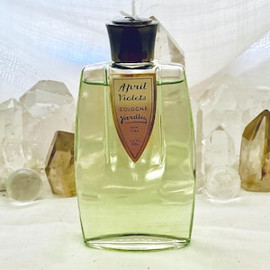 May include: A vintage glass bottle of "April Violets" cologne by Yardley. The bottle has a dark cap and a label with text. The liquid inside is light green. The bottle is surrounded by clear and amber crystals.
