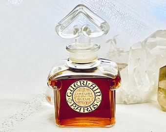 May include: A vintage perfume bottle with a clear, heart-shaped stopper. The bottle is filled with a deep amber liquid and features a gold label with the text "GUERLAIN L'HEURE BLEUE PARIS". The bottle has a square shape with rounded edges.