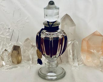 May include: A clear glass perfume bottle with a rounded, vertically ribbed body and a pedestal base. The bottle features a clear glass stopper, a black cord tied around the neck, and a small circular gold-colored tag with dark text.