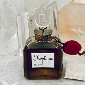 May include: A vintage glass bottle of perfume with a tulip-shaped stopper. The bottle is filled with a dark brown liquid and has a label that reads "Réplique Raphael Paris". The bottle is sitting on a white surface with a red wax seal and a white piece of paper.