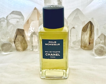 May include: A rectangular glass bottle of "Pour Monsieur" Eau de Toilette by Chanel. The bottle has a dark blue cap and a label with white text on a dark teal background. The liquid inside is yellow. The background features clear quartz crystals.