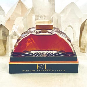 May include: A perfume bottle with a geometric design, filled with a reddish-brown liquid, sits on a black box with gold accents. The bottle is labeled "KL PARFUMS LAGERFELD - PARIS." The background features clear quartz crystals.