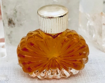 May include: A small, vintage perfume bottle with a silver-colored cap. The bottle is a warm amber color and has a textured, carved design. The bottle is filled with a liquid, and the design resembles a flower.