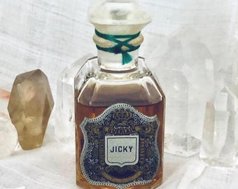 May include: A clear glass perfume bottle filled with amber liquid, featuring a faceted stopper tied with a green cord. The front label displays the text GUERLAIN and JICKY within an ornate, shield-shaped border with a crown graphic at the top.
