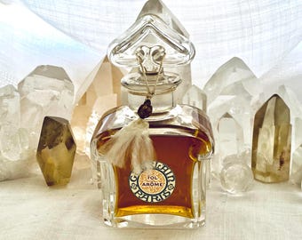 May include: A vintage perfume bottle with a clear glass stopper and a decorative tassel. The bottle is filled with amber-colored liquid and features a circular label with the text "Fol Arôme Paris". The background includes several clear and amber quartz crystals.