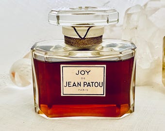 May include: A vintage perfume bottle of "JOY de JEAN PATOU" with a clear, faceted stopper. The bottle contains a deep red liquid and has a gold-coloured band around the neck. The label is white with black text. The bottle is displayed on a white surface.