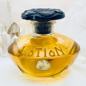 May include: A vintage amber-colored perfume bottle with a black floral stopper. The word "Lotion" is embossed on the front. A small, round emblem is on the front of the bottle. The bottle is filled with a golden liquid.