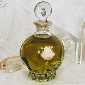 K&ouml;nnte beinhalten: Eine antike Glasflasche mit Parf&uuml;m, einem durchsichtigen Glasverschluss und einem goldenen Kronendesign. Die Flasche ist mit einer gr&uuml;nen Fl&uuml;ssigkeit gef&uuml;llt und tr&auml;gt ein Etikett mit der Aufschrift "Eau de Cologne".