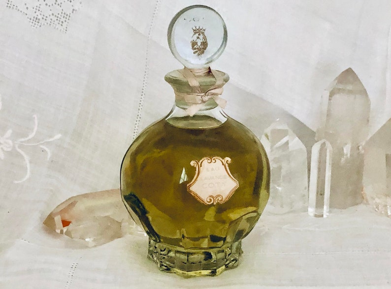 K&ouml;nnte beinhalten: Eine antike Glasflasche mit Parf&uuml;m, einem durchsichtigen Glasverschluss und einem goldenen Kronendesign. Die Flasche ist mit einer gr&uuml;nen Fl&uuml;ssigkeit gef&uuml;llt und tr&auml;gt ein Etikett mit der Aufschrift "Eau de Cologne".