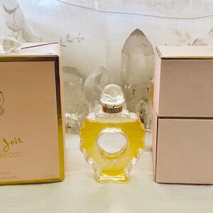 May include: Two pink boxes with gold trim, one containing a bottle of Coeur Joie Nina Ricci perfume. The bottle is clear glass with a gold top and a heart-shaped design.