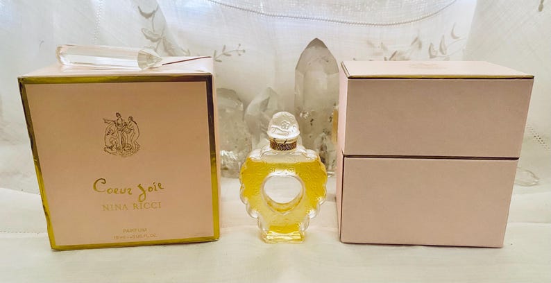 May include: Two pink boxes with gold trim, one containing a bottle of Coeur Joie Nina Ricci perfume. The bottle is clear glass with a gold top and a heart-shaped design.