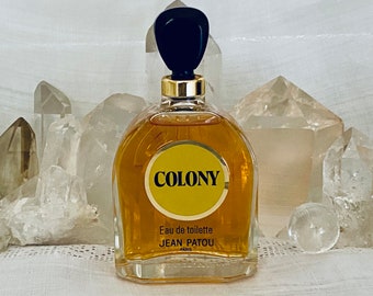 May include: A clear glass perfume bottle filled with amber liquid, featuring a black stopper and a gold collar. The front label is a yellow circle with the text COLONY, Eau de toilette, JEAN PATOU, PARIS printed in black lettering.