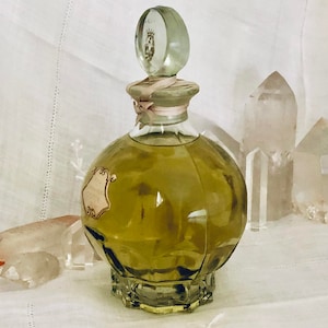 K&ouml;nnte beinhalten: Eine Vintage-Parf&uuml;m-Glasflasche mit einem durchsichtigen Glasstopfen und einem rosa Band. Die Flasche ist mit einer gr&uuml;nen Fl&uuml;ssigkeit gef&uuml;llt und hat ein Etikett mit goldenen Buchstaben. Die Flasche steht auf einer wei&szlig;en Stoffoberfl&auml;che mit klaren Quarzkristallen.