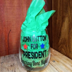 May include: A clear glass wine glass with a black vinyl sticker that reads "John Dutton for President, Ranching Done Right!" with a red star and a blue star on either side of the word "for".