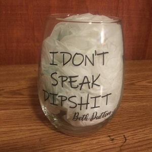 May include: A clear glass wine glass with the text "I don't speak dipshit" and the signature "Beth Dutton" printed on the side.