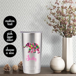 May include: A stainless steel tumbler with a floral elephant design and the name "Shelby" in pink lettering. The tumbler has a black lid and the word "YETI" printed on the bottom.