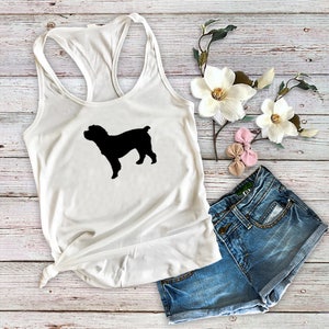 May include: White racerback tank top with a black silhouette of a dog. The tank top is paired with denim shorts, and decorative flowers. The dog silhouette is centered on the front of the tank top.