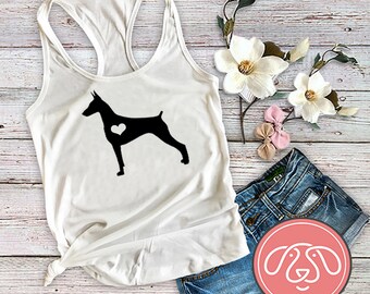 Pode incluir: Camisola branca de alças com uma silhueta preta de um Doberman Pinscher com um coração branco no peito. A camisola está sobre uma superfície de madeira com um par de calções de ganga azul e flores.