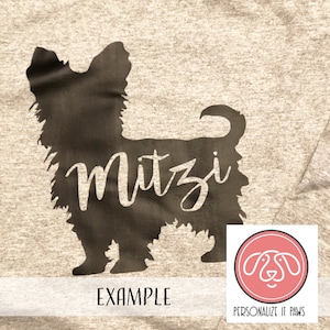 May include: A brown silhouette of a Yorkshire Terrier with the name "Mitzi" written inside the silhouette. The image is on a light brown t-shirt with the text "EXAMPLE" below the silhouette and a logo for "PERSONALIZE IT PAWS" in the bottom right corner.