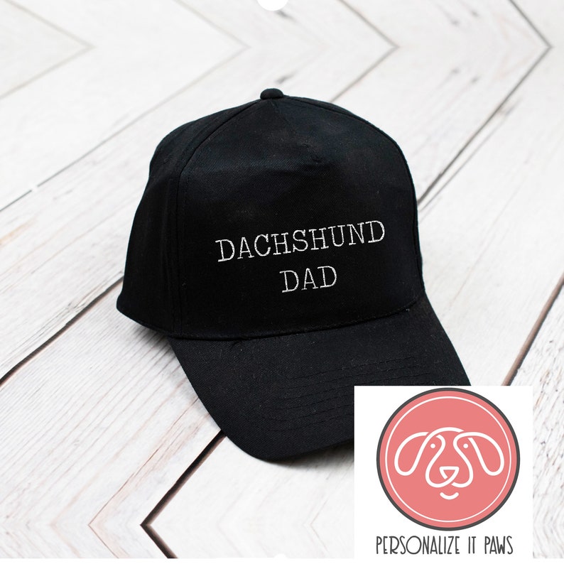 May include: Black baseball cap with white embroidered text "Dachshund Dad". The cap has a curved brim and a white logo with a dog silhouette on the right side of the image. The logo says "Personalize It Paws".