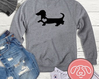 Könnte beinhalten: Ein graues Sweatshirt mit einer schwarzen Silhouette eines Dackels mit einem Herzen auf der Brust. Das Sweatshirt liegt auf einem weißen Holzgrund mit einer blauen Jeans, einer grauen Mütze und weißen Turnschuhen.