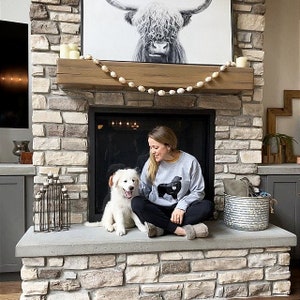 K&ouml;nnte beinhalten: Eine Frau sitzt auf einem steinernen Herd mit einem wei&szlig;en Hund. Sie tr&auml;gt ein graues Sweatshirt mit einer schwarzen Silhouette eines Hundes. Der Kamin hat einen Holzmantel mit einer Girlande aus wei&szlig;en Perlen.