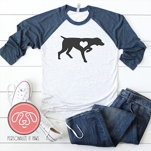 May include: A white and navy blue baseball tee shirt with a black silhouette of a German Shorthaired Pointer with a heart in its chest. The shirt is laying on a white wooden surface with a pair of white sneakers and blue jeans.