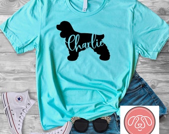May include: A turquoise t-shirt with a black silhouette of a cocker spaniel and the name "Charlie" printed on it. The shirt is laying on a wooden surface with a pair of white sneakers, blue denim shorts, and sunglasses.