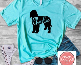 Puede incluir: Una camiseta turquesa con una silueta negra de un perro y el nombre "charlie" impreso en la parte delantera. La camiseta está sobre una superficie de madera con un par de zapatillas blancas, unas gafas de sol y unos pantalones cortos de mezclilla.