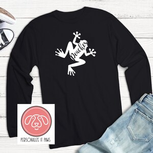 May include: Black long-sleeve shirt with a white frog graphic and the text "Peaches" on the front. The shirt is laid flat on a white wooden surface with a pair of black and white sneakers and blue jeans in the background.