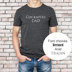 Könnte beinhalten: Ein dunkelgraues T-Shirt mit dem Aufdruck "COCKAPOO DAD" in Weiß. Das Shirt wird von einer Person getragen, die vor einer Backsteinwand steht. Schriftarten zur Auswahl: Bernard, Ariel, Trajan.