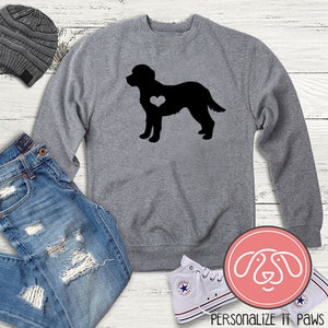 Op de afbeelding: Grijs sweatshirt met een zwart silhouet van een hond met een hart op zijn borst. Het sweatshirt ligt op een wit houten oppervlak met een spijkerbroek, een beanie en witte sneakers.