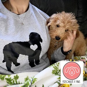 May include: A gray sweatshirt with a black silhouette of a dog with a heart on its chest. The sweatshirt is being worn by a person holding a small brown dog. The text "PERSONALIZE IT PAWS" is on a pink circle with a white outline.