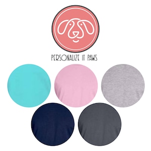 K&ouml;nnte beinhalten: F&uuml;nf T-Shirts in verschiedenen Farben mit einer wei&szlig;en Umrandung eines Hundekopfes in einem Kreis auf rosa Hintergrund mit dem Text "PERSONALIZE IT PAWS" darunter.