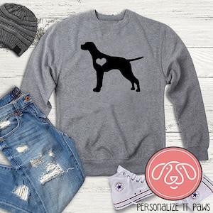 Op de afbeelding: Grijs sweatshirt met een zwart silhouet van een Duitse Korthaar met een hart op zijn borst. Het sweatshirt ligt op een wit houten oppervlak met een spijkerbroek, een beanie en witte sneakers.