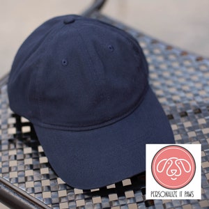 May include: A plain dark blue baseball cap with a curved brim. The cap is sitting on a black and white checkered surface. The text "PERSONALIZE IT PAWS" is on a white label with a pink paw print.