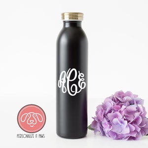 Op de afbeelding: Zwarte roestvrijstalen waterfles met een gouden deksel en een wit monogram "RPE" aan de voorkant. De fles staat op een wit oppervlak naast een bos paarse bloemen.