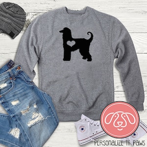 Peut inclure: Sweat-shirt gris avec une silhouette noire d'un lévrier afghan avec un cœur sur la poitrine. Le sweat-shirt est posé sur une surface en bois blanc avec un jean bleu, un bonnet gris et des baskets blanches.