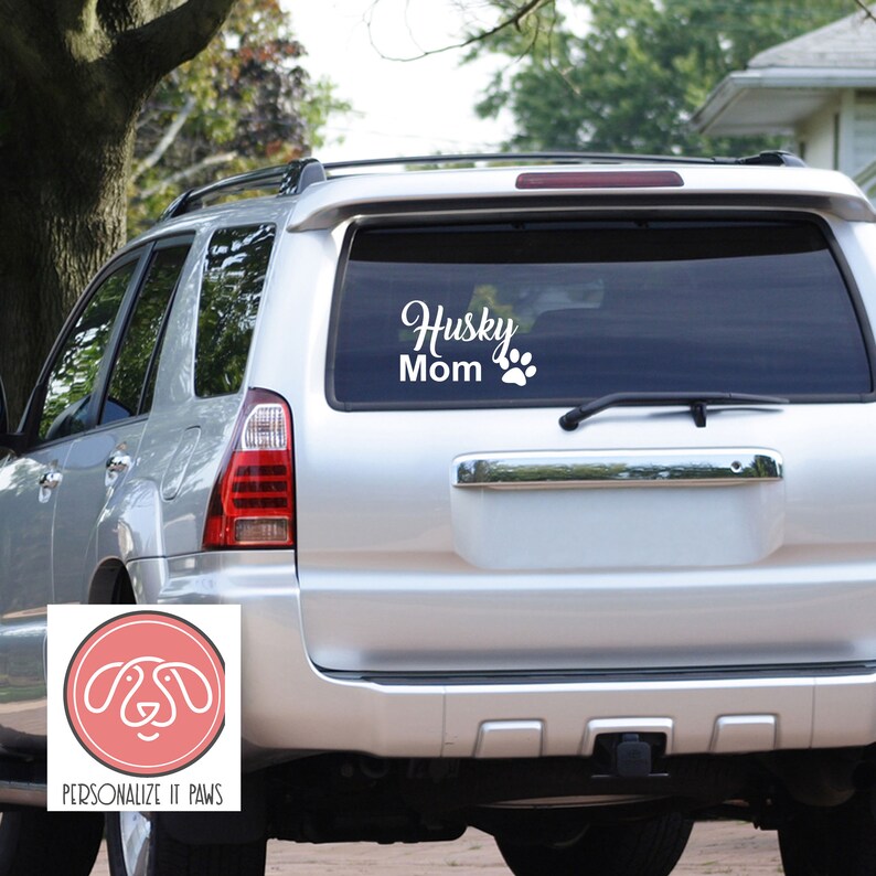 Pu&ograve; includere: Un'auto bianca con un adesivo sul lunotto posteriore che dice "Husky Mom" con un'impronta di zampa. L'adesivo &egrave; su un'auto argentata con un paraurti nero.