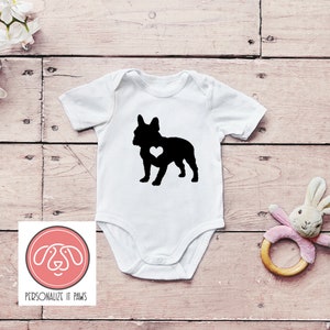 May include: White baby onesie featuring a black silhouette of a French Bulldog with a white heart on its chest. The onesie is displayed against a wooden backdrop.