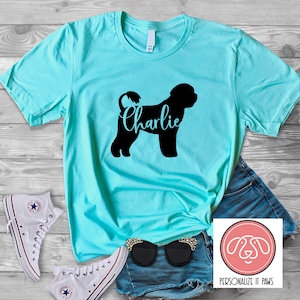 May include: A turquoise t-shirt with a black silhouette of a Portuguese Water Dog and the name "Charlie" printed on it. The shirt is laying on a wooden surface with a pair of white sneakers, blue denim shorts, and sunglasses.