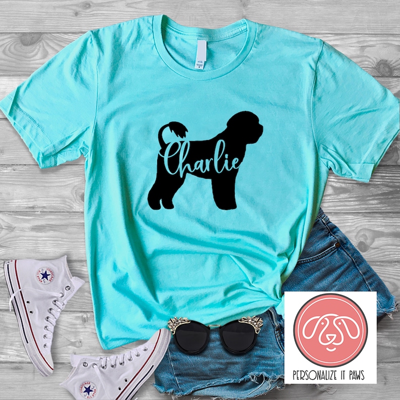 May include: A turquoise t-shirt with a black silhouette of a Portuguese Water Dog and the name "Charlie" printed on it. The shirt is laying on a wooden surface with a pair of white sneakers, blue denim shorts, and sunglasses.